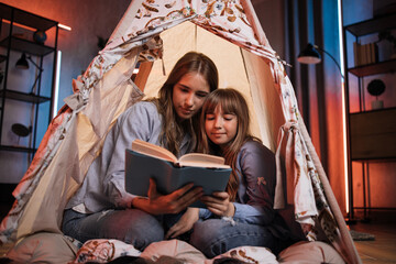 Caucasian pretty blond sisters sitting in wigwam and reading book during evening time. Cozy home and family concept. © sofiko14