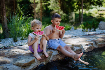 Little chidren sitting near lake and eating watermelon on hot sunny day during summer vacation.