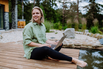 Woman, freelancer working on laptop, sitting on a pier by the backyard lake, a concept of remote...