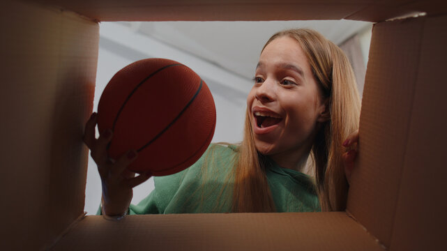 Happy Surprised Winner Girl Unpacking Delivery Parcel At Home. Smiling Satisfied Young Child Kid Shopper Online Shop Customer Opening Cardboard Box Receiving Purchase Gift Ball By Fast Postal Shipping