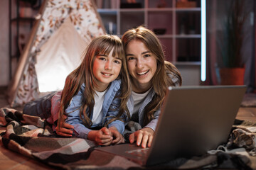 Beautiful cute sisters or friends lying on floor embrace outside wigwam using laptop computer. Happy girls spending evening time together watching favorite movie on digital device at home.
