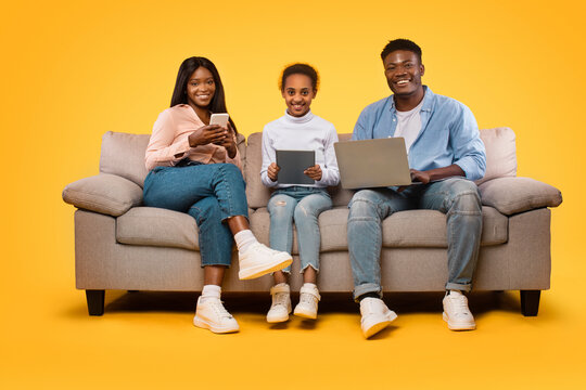 Technologies for family. Happy black mom, dad and their daughter sitting with different gadgets, yellow background