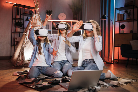 Happy mother and cute daughters sitting near toy wigwam at home and using modern VR headset during evening time. Caucasian family using modern technologies for playing at home.