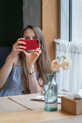 Young woman sits at table in cafe and takes selfie. Girl in coffee shop with phone in her hands. Vertical frame.