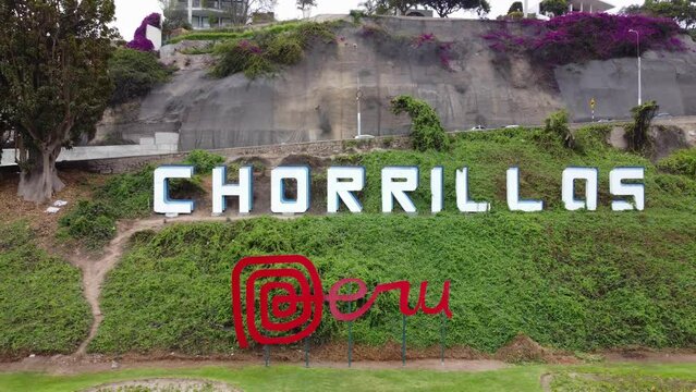 Drone video of a red sign reading "Peru" and behind it on a green hill, another sign reading "Chorrillos" the name of the district in Lima,Peru. Drone slowly elevates and flies backwards.