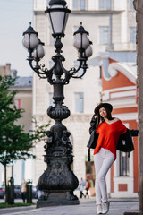 Fototapeta premium Vertical shoot of standing attractive brunette woman in hat, leather jacket and white pants over old city on background. Hispanic female model exploring world on vacations. Leisure and travel.