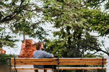 A young long-haired, bearded man sits in a park on a bench outdoors on the green trees, enjoying...