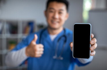 Smiling young asian nurse in uniform with stethoscope shows smartphone with blank screen and thumb up