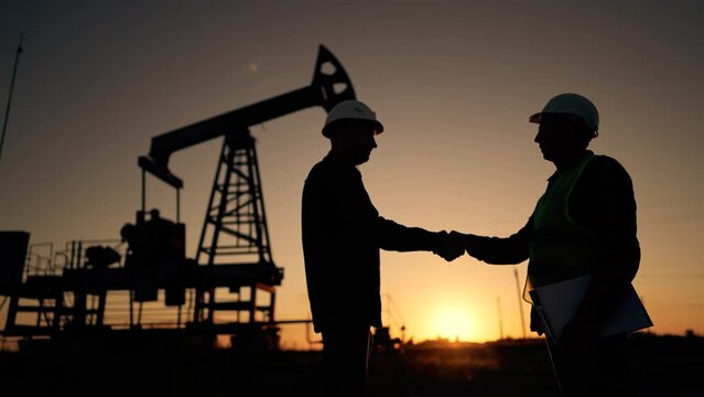 Teamwork.Oil engineer in helmet work with an oil pump.Silhouette of worker in field.Oil pump rig work at sunset.Two businessmen sign contract with handshake.Oil business concept.Work mineral deposit