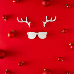 Christmas deer concept. Creative layout made of reindeer antlers, hipster glasses and Christmas balls on red background. Minimal flat lay. New year holiday idea. Greeting card.