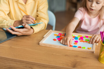 Professional teacher and pupil girl exercising with letters, sitting at table, making various words on board