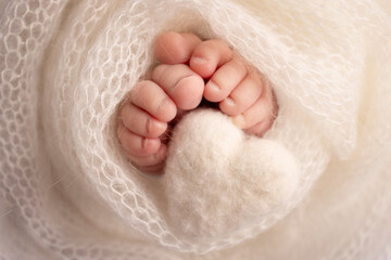 Knitted white heart in the legs of a baby. Soft feet of a new born in a white wool blanket. Close-up of toes, heels and feet of a newborn. Macro photography the tiny foot of a newborn baby.