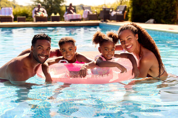 Portrait Family On Summer Holiday With Two Girls Floating In Swimming Pool On Inflatable Ring
