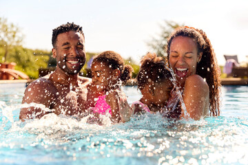 Family On Summer Holiday With Two Girls Being Held In Swimming Pool By Parents And Splashing - obrazy, fototapety, plakaty