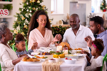 Multi-Generation Family Celebrating Christmas At Home Saying Prayer Before Eating Meal Together