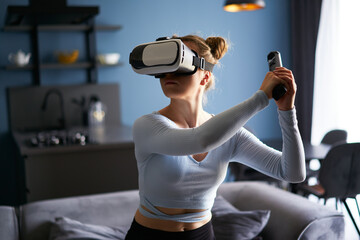 Young futuristic blonde girl wearing virtual reality headset, holding controller playing a video...