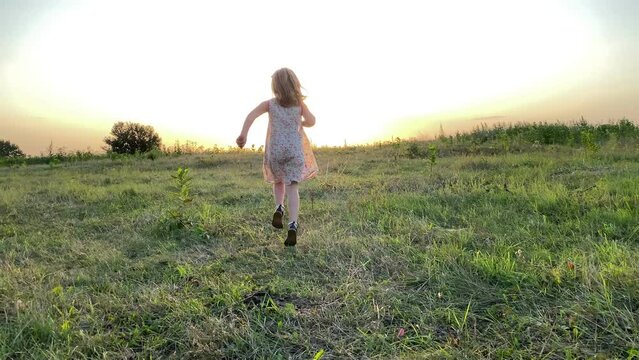 Rear view of cheerful little girl running skipping on meadow forward golden lights of summer sunset. Lovely female kid having fun at countryside. Lovely child enjoying pastime at nature. Simple living