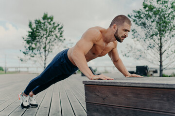Obraz premium Confident muscled sportsman in sport wear doing push up outdoors, works on biceps and trains determination. Strong topless athletic man exercises in open air. People, motivation, training concept