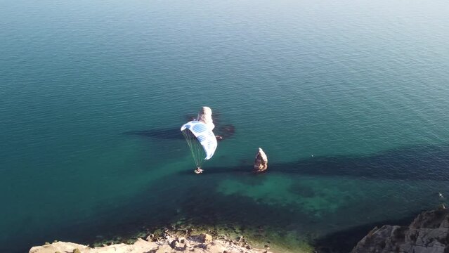 Aerial drone view of a man flying a white and blue paraglider over a hill and trees to the sea waves near the rocks. Active paraglider flight over the seascape with clear skies at suset. Extreme sport