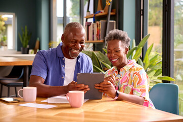 Smiling Senior Couple Sitting Around Table At Home Reviewing Finances Using Digital Tablet