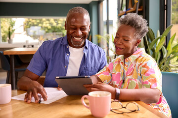 Smiling Senior Couple Sitting Around Table At Home Reviewing Finances Using Digital Tablet - Powered by Adobe