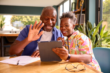 Smiling Senior Couple Sitting Around Table At Home Making Video Call On Digital Tablet