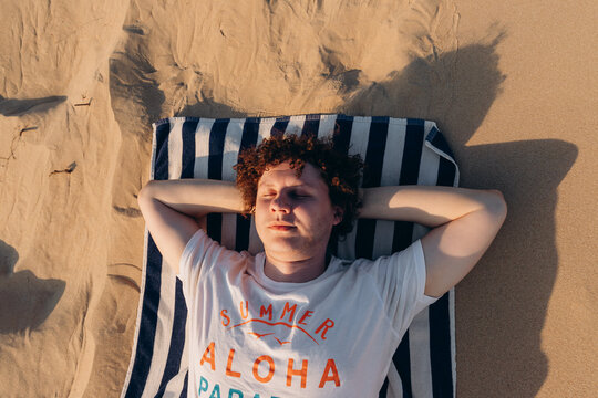 Young Caucasian man with curly hair, closed eyes, resting, relaxing or sleeping in white T-shirt with words Summer Aloha lies on striped towel on sand beach barkhan or sand dune at sunset. Portrait