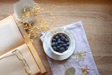 Vintage porcelain cup filled with fresh blueberries, pressed flowers, open book, reading glasses and lit candle on the table. Hygge at home. Top view.