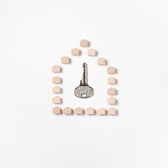 Wooden figures of houses are stacked in a closed ring in the form of a building, and in the center there is a metal key on a light gray textural background in a square orientation.