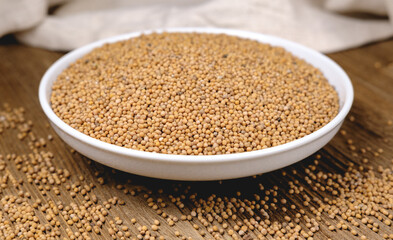 Mustard seeds are poured into a white bowl, standing on a wooden table against a background of light textured and wrinkled fabric from a storage bag.