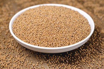 Mustard seeds are poured into a white round bowl standing on a textured wooden table.