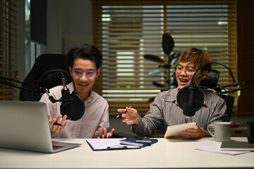 Two happy man using laptop and microphone streaming audio podcast at home studio. Entertainment,...
