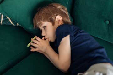 Portrait of serious, scary, sad and depressed little boy lying on green sofa, sucking thumb finger....