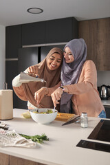 Two muslim friends smiling while pouring olive oil and tossing a salad at modern kitchen.