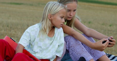 Landscape of straw bales against setting sun on background. Sisters watch video on smartphone outdoors. Childhood. Country life