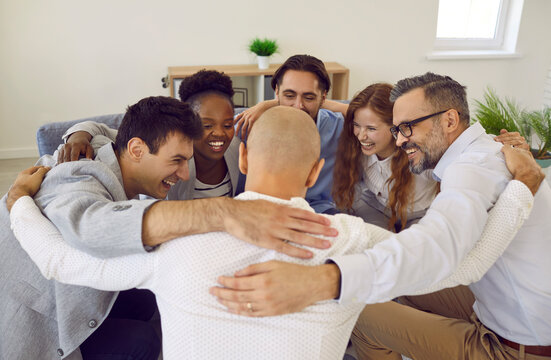 Team of successful positive colleagues laughing and hugging while standing in circle in office. Cheerful business colleagues celebrate their success together. Concept of cohesion and teamwork.