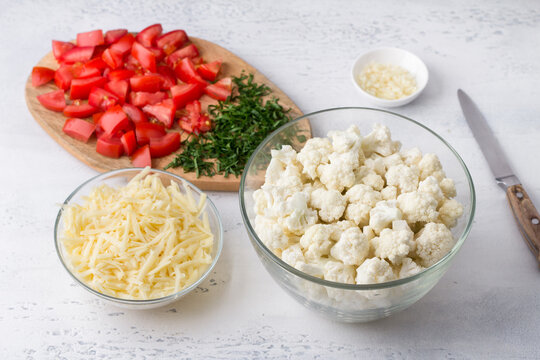 Ingredients for cooking cauliflower casserole: cauliflower cut into florets, grated cheese, chopped tomatoes and parsley, chopped garlic on a light blue background