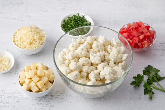 Ingredients for cooking cauliflower casserole: cauliflower cut into florets, grated cheese, chopped tomatoes and parsley, chopped garlic on a light blue background