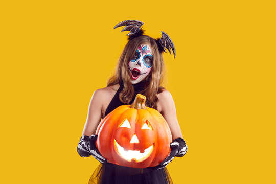Portrait of child in creative Halloween costume. Girl with skull makeup standing isolated on yellow background, holding Jack o lantern pumpkin and looking at camera with surprised face expression