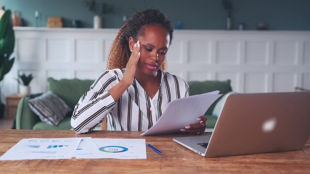 Upset young African American woman accountant gets angry and clutches head when sees error in financial documents or audit reports sits at table with laptop in home office. Crisis management concept