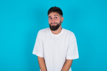Portrait of mysterious bearded caucasian man wearing white T-shirt over blue background looking up with enigmatic smile. Advertisement concept.
