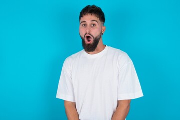 Obraz premium Expressive facial expressions. Shocked stupefied bearded caucasian man wearing white T-shirt over blue background, keeps jaw dropped feels stunned from what he sees aside.