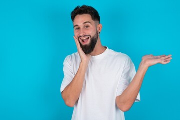 Positive glad bearded caucasian man wearing white T-shirt over blue background says: wow how exciting it is, indicates something.  One hand on his head and pointing with other hand.