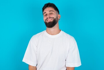 Obraz premium bearded caucasian man wearing white T-shirt over blue background with broad smile, shows white teeth, feeling confident rejoices having day off.