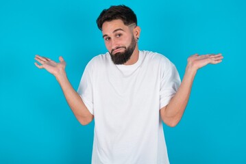 Fototapeta premium Puzzled and clueless bearded caucasian man wearing white T-shirt over blue background with arms out, shrugging shoulders, saying: who cares, so what, I don't know. Negative human emotions.