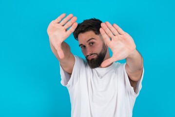 Portrait of smiling bearded caucasian man wearing white T-shirt over blue background looking at camera and gesturing finger frame. Creativity and photography concept.