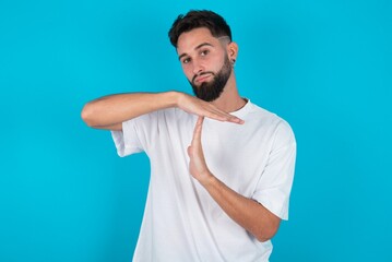 Fototapeta premium bearded caucasian man wearing white T-shirt over blue background feels tired and bored, making a timeout gesture, needs to stop because of work stress, time concept.