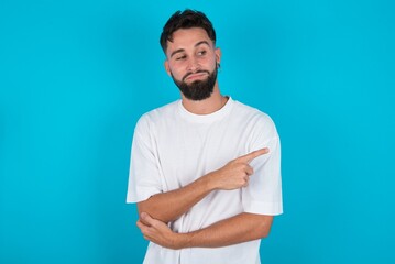 Portrait of bearded caucasian man wearing white T-shirt over blue background posing on camera with tricky look, presenting product with index finger. Advertisement concept.