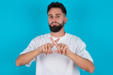 bearded caucasian man wearing white T-shirt over blue background has rejection angry expression...