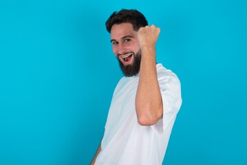 Naklejka premium Overjoyed bearded caucasian man wearing white T-shirt over blue background glad to receive good news, clenching fist and making winning gesture.